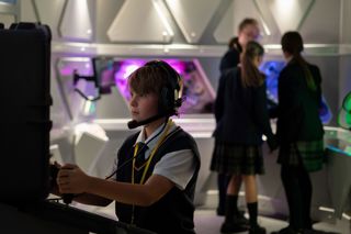 A child with headphones and a microphone on a computer controlling a Mars Rover. Other children are talking in the background.