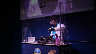 An Education Presenter in a lab uniform, behind a perspex screen, performing a science experiment.