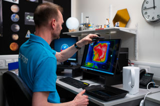 An Education Presenter holding up a thermal imaging camera above some boiled water in a tray, on a Video Conference to a School Group.