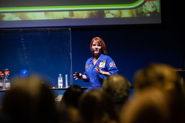 An Education Presenter is on stage in a blue flight suit, presenting a talk to a public audience.